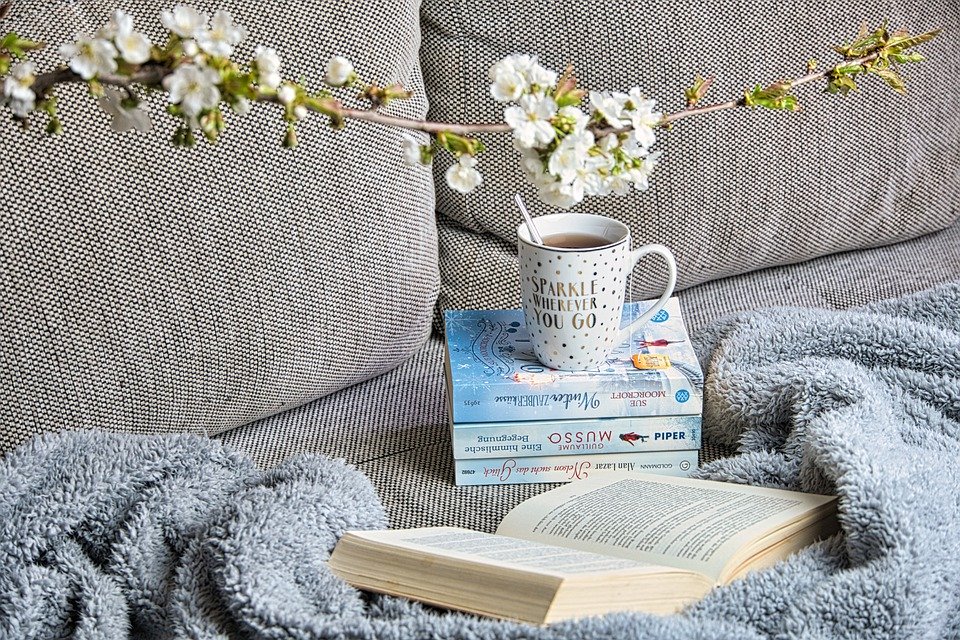 Auf einem gemütlichen Sofa liegt eine Decke und mehrere Bücher, wobei auf einem davon eine Teetasse steht